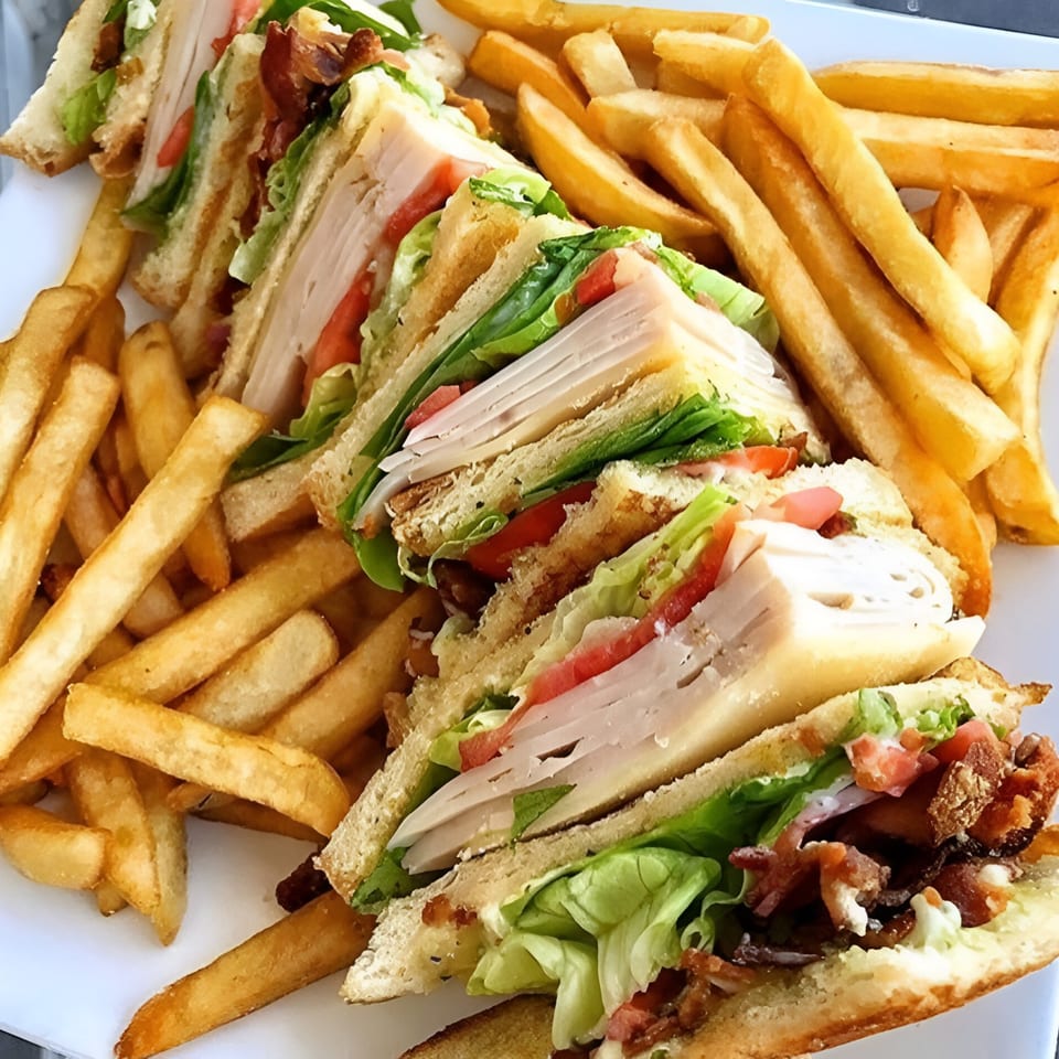 A large club sandwich, cut into quarters, with turkey, bacon, lettuce, and tomato, served with fries.